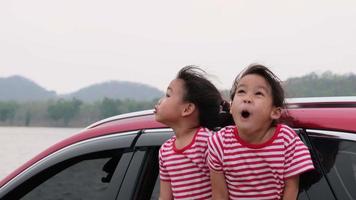 Cute Asian siblings girls smiling and having fun traveling by car and looking out of the car window. Happy family enjoying road trip on summer vacation.