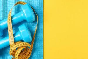 Fitness concept. Dumbbells and measuring tape on a blue and yellow background, top view flat lay