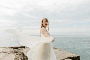 A young beautiful girl in a long milk-colored dress walks along the beach and pier against the background of the sea. photo