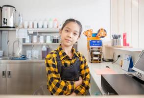 Asian little girl wearing apron happy face smiling with crossed arms, learning cashier operating at the cash desk in cafe, education concept photo