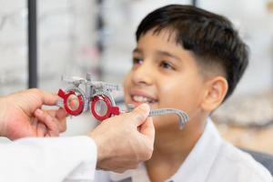 Selective focus at trial frame, Smiling Indian boy choosing glasses in optics store, Boy doing eye test checking examination with optometrist using trial frame in optical shop photo