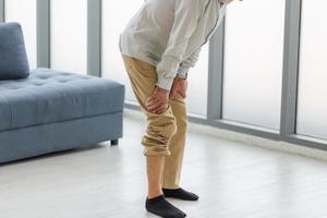 Old man suffering from knee pain in the living room, Elderly man suffering from knee pain while standing in living room photo