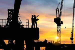 silueta de ingeniero y trabajador revisando el proyecto en el fondo del sitio de construcción de infraestructura pesada, sitio de construcción al atardecer por la noche. foto