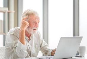Portrait of Worried senior man using laptop and smartphone at home, Mature man in living room with laptop browsing internet on modern computer gadget photo