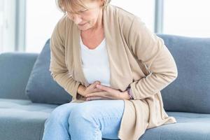 Unhappy woman stomach ache, mature woman with stomach pain feeling unwell sitting in living room photo