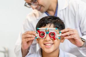 niño indio-tailandés sonriente eligiendo anteojos en la tienda de óptica, niño haciendo un examen de control de la vista con un optometrista usando un marco de prueba en la tienda de óptica foto