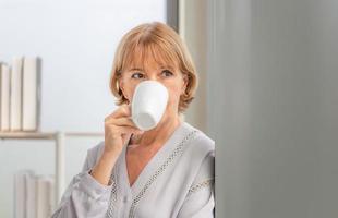 Senior woman inside new home during a coffee break, Woman standing near window with cups of coffee photo