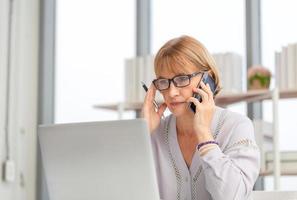 Portrait of woman using laptop and smartphone at home, Mature woman in living room with laptop browsing internet on modern computer gadget photo