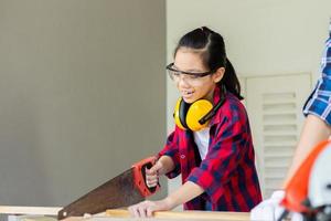 Kid learning woodworking in the craftsman workshop, Asian girl standing with noise reduction earmuffs in a carpentry workshop, Manual wood worker carpenter with saw photo