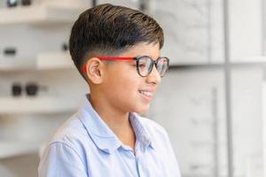 niño indio sonriente eligiendo anteojos en la tienda de óptica, retrato de un niño de raza mixta que usa anteojos en la tienda de óptica