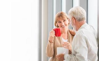 pareja de ancianos hablando mientras está de pie cerca de la ventana con tazas de café, pareja de ancianos dentro de un nuevo hogar durante la pausa para el café foto