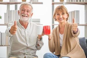 Senior couple inside home during a coffee break, Smiling elderly couple holding cups of coffee with showing thumbs up photo