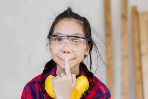 Chica asiática de pie con orejeras de reducción de ruido hace cara graciosa en un taller de carpintería foto