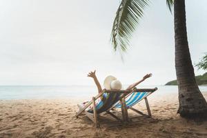 Una joven asiática relajándose en una silla de playa le levanta la mano con un sombrero flexible foto