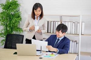 An Asian female coworker stood talking to a business man on the desk in her hand holding a cup of coffee. photo