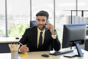 A business man in a suit that is neatly dressed sitting on the phone with a bright smile in the office photo