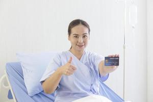 una paciente asiática yacía en la cama del hospital en su mano, tenía una tarjeta de crédito y los pulgares hacia arriba para mostrar una sonrisa. foto