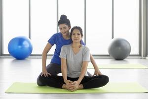 A female trainer is teaching a middle-aged Asian female customer to do yoga on a rubber mat in a gymnasium. photo