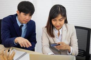 A young professional Asian businesswoman who uses a laptop computer and a credit card to pay online while sitting at her desk, with a male friend next to him.
