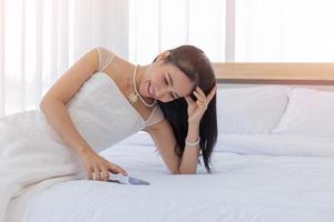 una novia asiática con un vestido de novia blanco acostada en la cama tocando su teléfono con una sonrisa bastante buena. foto