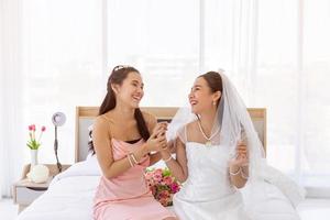 Asian brides in a white wedding dress and brides in a pink dress sitting on the bed together smiling brightly. photo