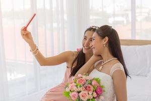 The bride's friend is using a smartphone to take a selfie of an Asian bride holding a bouquet of beautiful flowers in a wedding dress in the dressing room on the bed. photo