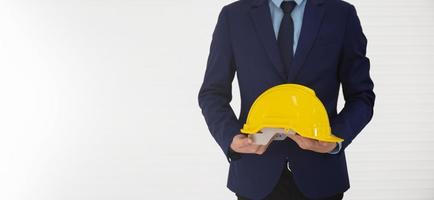 A businessman stands and holds a yellow helmet in his hand and behind him is a white wall. photo