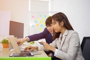 A young professional Asian businesswoman who uses a laptop computer and a credit card to pay online while sitting at her desk, with a male friend next to him. photo