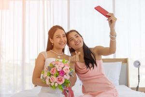 The bride's friend is using a smartphone to take a selfie of an Asian bride holding a bouquet of beautiful flowers in a wedding dress in the dressing room on the bed and making a mini heart hand sign.