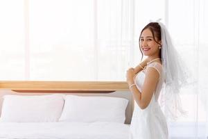 una novia asiática vestida con un vestido de novia blanco se para con una linda y brillante sonrisa al lado de la cama en la habitación.