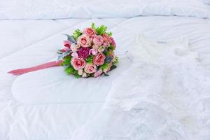 A beautiful bouquet of flowers was laid out on a clean, white bed. Next to the bridal gown were placed together. photo