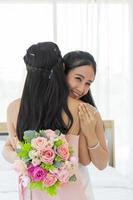 una novia asiática con un vestido de novia blanco abraza a su amiga con un vestido rosa con una sonrisa, luego muestra un anillo de diamantes en su dedo izquierdo y, por otro lado, sostiene un hermoso ramo de flores. foto