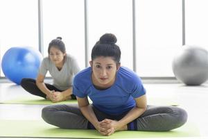 Two middle-aged Asian women doing yoga sitting on a rubber mat in a gym. photo