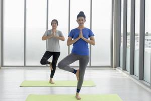 Two middle-aged Asian women doing yoga standing on rubber mats in the gym. photo
