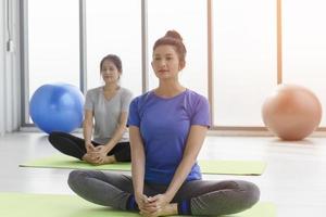 Two middle-aged Asian women doing yoga sitting on a rubber mat in a gym. photo