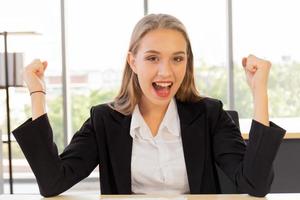 A beautiful business woman in a suit, neatly dressed, sitting in the office, excitedly happy, raised his hand with a bright smile on the table photo