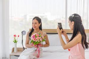 The bride's friend is using a smartphone to take a photo of an Asian bride in a wedding dress in her hand, holding a large, beautiful bouquet of flowers sitting on the bed.