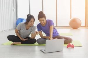 Two middle-aged Asian women are looking at how to do basic yoga from their laptop in the gym. photo