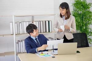 An Asian female coworker stood talking to a business man on the desk in her hand holding a cup of coffee. photo