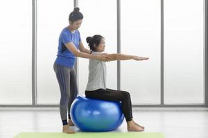 una entrenadora le está enseñando a una clienta asiática de mediana edad a hacer yoga sentada en una pelota de goma en un gimnasio.