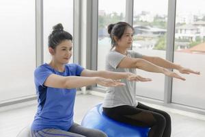 dos mujeres asiáticas de mediana edad haciendo yoga sentadas en una pelota de goma en el gimnasio. foto