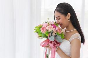 An Asian bride in a wedding dress is sitting smiling brightly on the bed in her hand holding a beautiful bouquet of flowers. photo