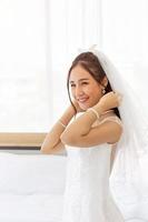 An Asian bride dressed in a white wedding dress stands in a cute and bright smile beside the bed in the room. photo