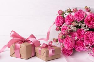 rosas frescas de color rosa pastel y cajas de regalo envueltas en papel kraft con cintas sobre una mesa de madera blanca. foto