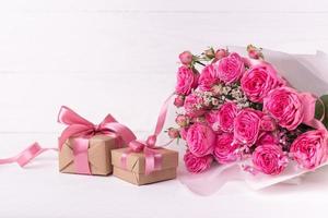 rosas frescas de color rosa pastel y cajas de regalo envueltas en papel kraft con cintas sobre una mesa de madera blanca.