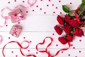 Red ribbon shaped as heart, bouquet of roses, hearts and gift boxes wrapped in kraft papper on white wooden table.