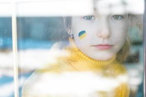 A sad child look at the window with the flag of Ukraine painted on the cheek, worries and fear. Humanitarian aid to children, world peace, security. photo