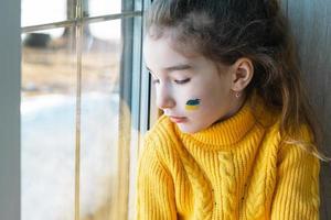 un niño triste mira la ventana con la bandera de ucrania pintada en la mejilla, preocupaciones y miedo. ayuda humanitaria a los niños, paz mundial, seguridad. foto