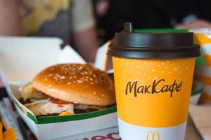 A paper cup of McDonald's coffee with the inscription Maccafe in Russian and a hamburger in a box on a tray. Fast food restaurant chains. Russia, Kaluga, March 21, 2022.
