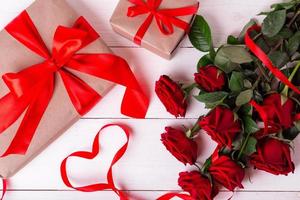 Red ribbon shaped as heart, bouquet of roses and gift boxes wrapped in kraft papper on white wooden table. photo